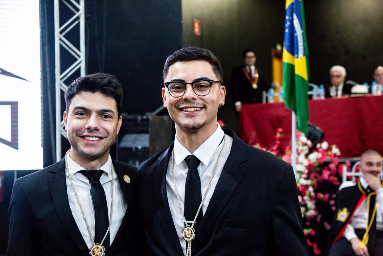Dois alumni abraçados e sorrindo, de terno e com o colar da ADASP no peito, durante cerimônia