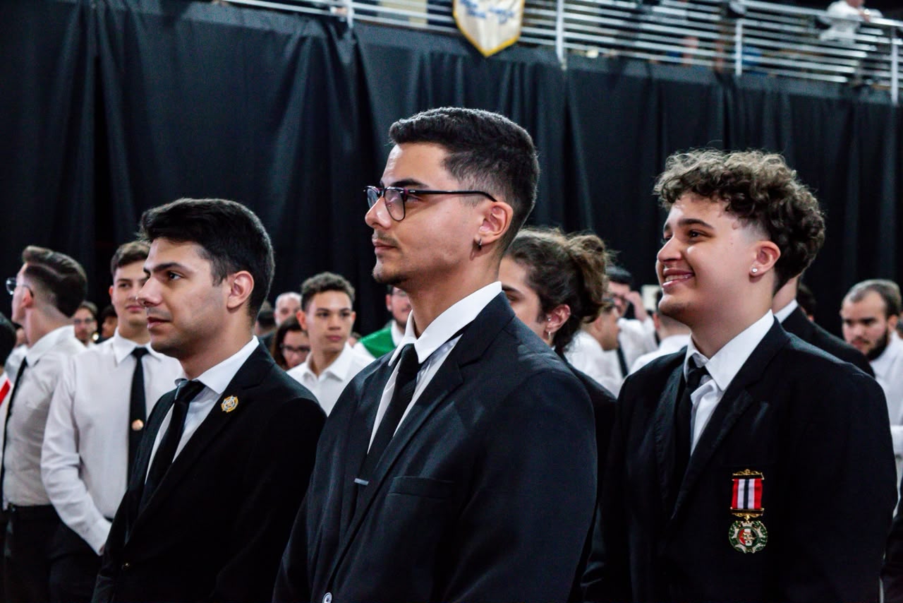 Três alumni lado a lado durante cerimônia; um deles sorri, com a medalha nas cores de São Paulo no peito